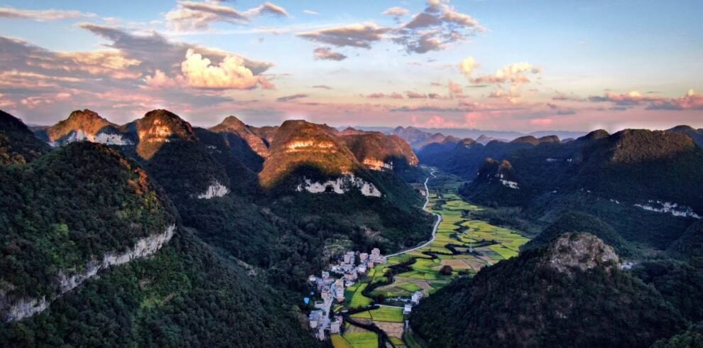 guizhou, chinatrip, A winding river or road through a lush valley surrounded by towering forested karst mountains at sunset, with a small village at the base of the hills.