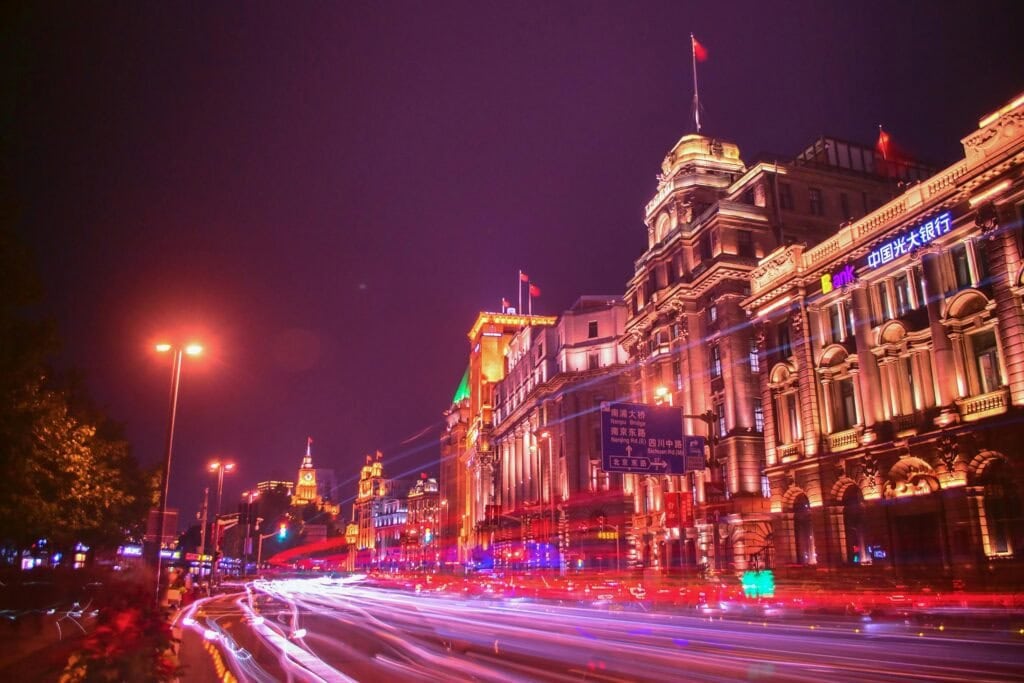 shanghai, chinatrip, Nighttime city street with ornate, illuminated historic buildings and red/white light trails from moving vehicles along the curbside lanes.