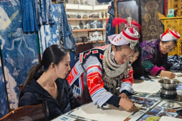 wax painting, guizhou, chinatrip, A woman in a black jacket sits at a table with a group of people in traditional dress, examining papers in a crafts shop filled with textiles and cultural artifacts.