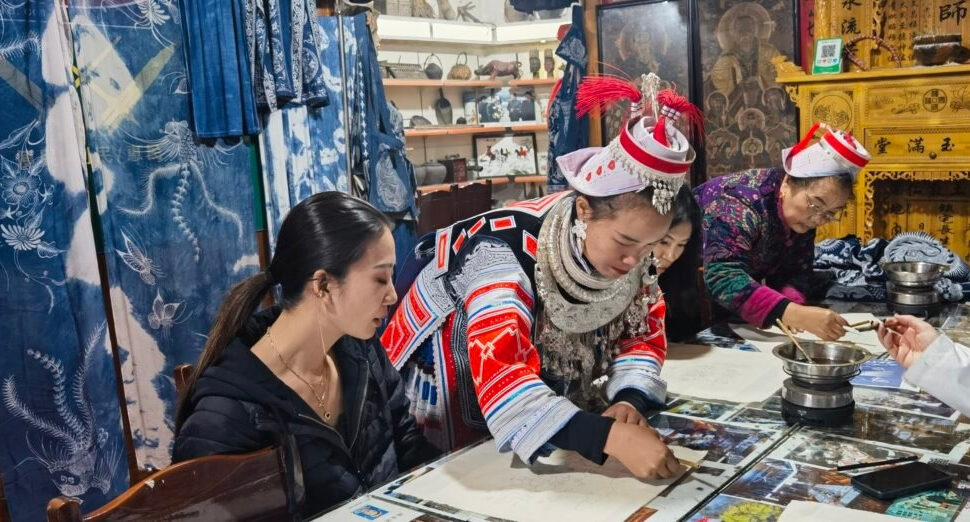 wax painting, guizhou, chinatrip, A woman in a black jacket sits at a table with a group of people in traditional dress, examining papers in a crafts shop filled with textiles and cultural artifacts.
