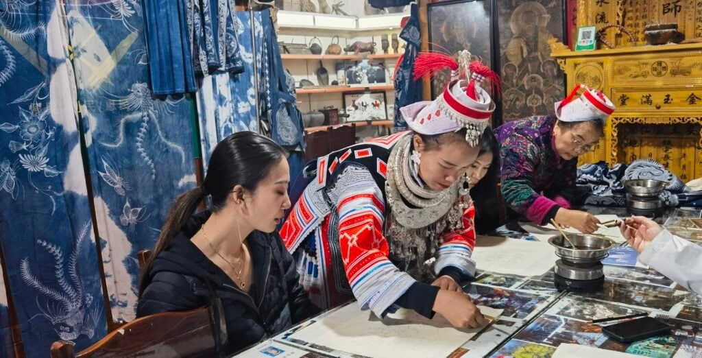 wax painting, guizhou, chinatrip, A woman in a black jacket sits at a table with a group of people in traditional dress, examining papers in a crafts shop filled with textiles and cultural artifacts.