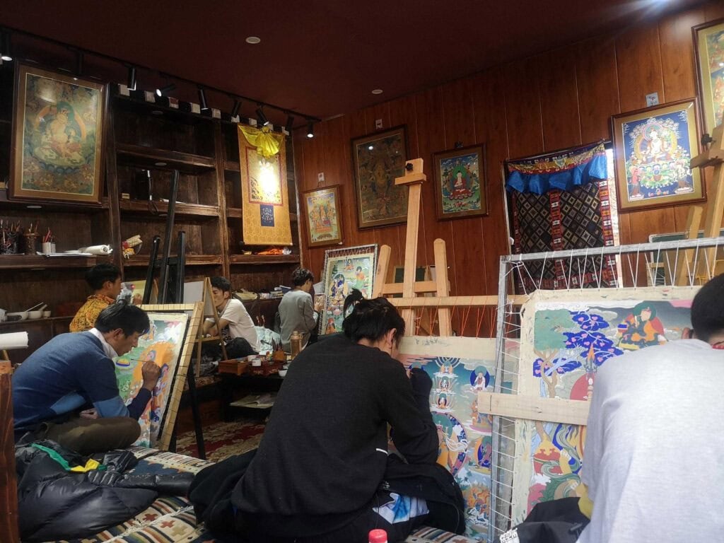 tangka, tibet, chinatrip, Art studio with several people painting on large colorful canvases, surrounded by wooden walls, framed artworks, and easels; artists are seated on the floor or on cushions working intently.
