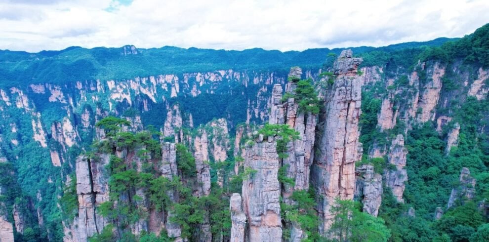 zhangjiajie, hunan, chinatrip, Towering sandstone pillars rise from a dense green forest under a cloudy blue sky.