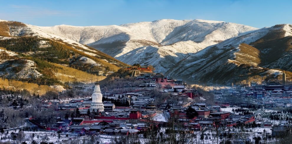 wutai, chinatrip, Snow-covered mountain village with a white stupa-like tower in the foreground and red-roofed buildings scattered across the valley.