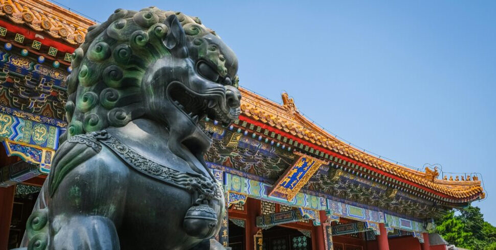 the forbidden city, beijing, chinatrip, Bronze guardian lion statue in front of a colorful Chinese temple with ornate roof details.