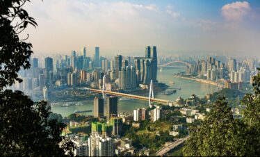 Panoramic cityscape with high-rise buildings along a winding river, framed by green hills and trees in the foreground. Two bridges span the river among dense skyscrapers.",