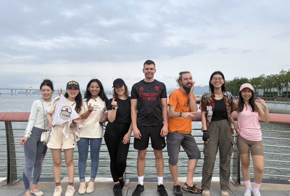 china explorer tour team, chinatrip, Group of eight friends posing on a waterfront boardwalk with railing and cloudy sky behind them.