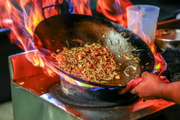 Traditional Chinese cuisine, Stir-fried noodles sizzling in a wok over a blazing flame, as a cook tilts the pan with a hand visible on the edge.