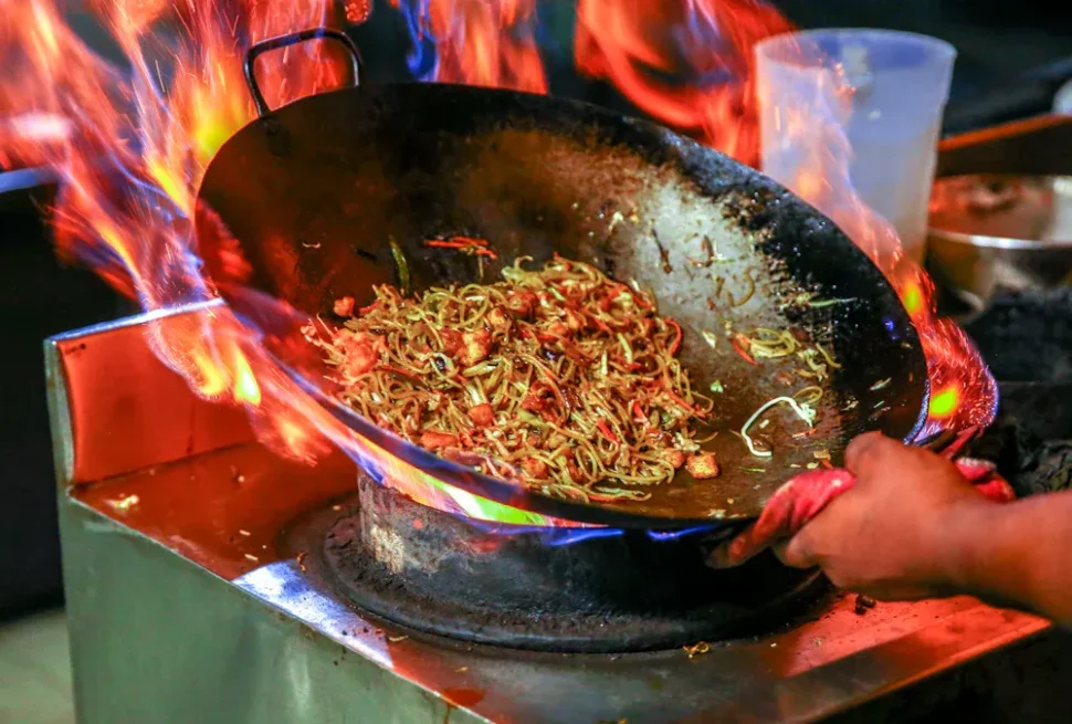 Traditional Chinese cuisine, Stir-fried noodles sizzling in a wok over a blazing flame, as a cook tilts the pan with a hand visible on the edge.
