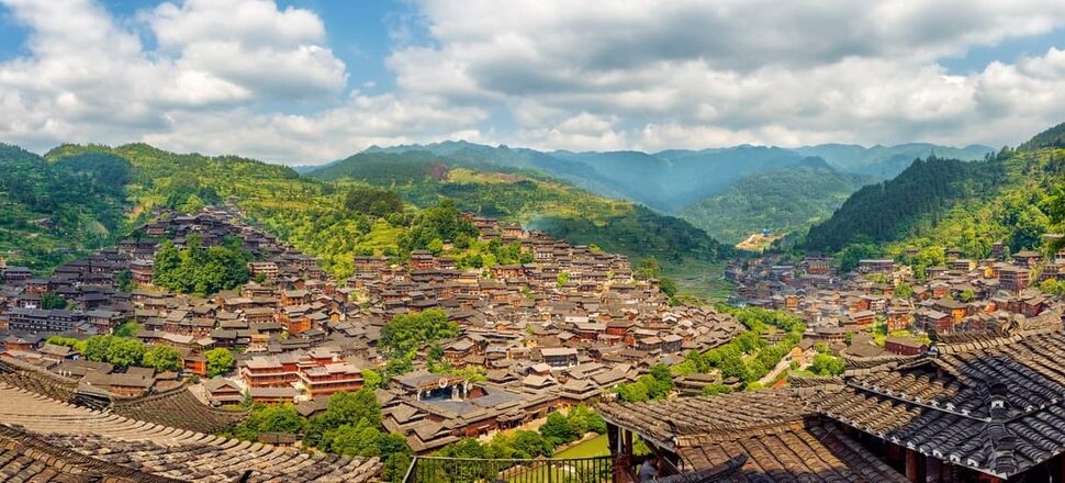 Panoramic view of a hillside village with densely packed wooden buildings and tiled roofs amid green mountains.