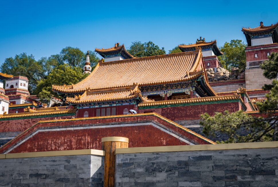Traditional Chinese temple complex with ornate yellow-tiled roofs, red walls, and blue sky background on a hillside area of trees and stone walls.