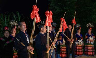 School trip in Guizhou, chinatrip, Group of people in traditional clothing holding tall bamboo poles with red fabric bows at a nighttime festival.