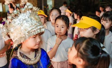 School trip in Guizhou, chinatrip, Children in a crowded festival, a girl in blue traditional dress with a beaded headdress among peers wearing yellow caps and white shirts.