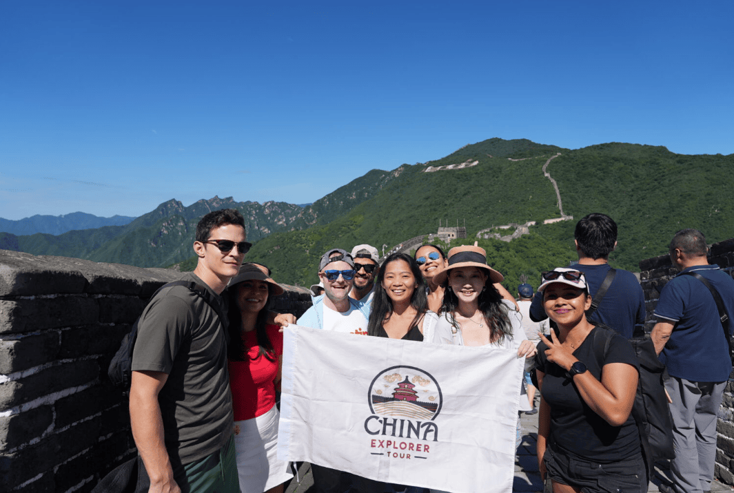 Group of travelers posing with a China Explorer Tour banner on the Great Wall, blue sky above and green mountains in the background.",