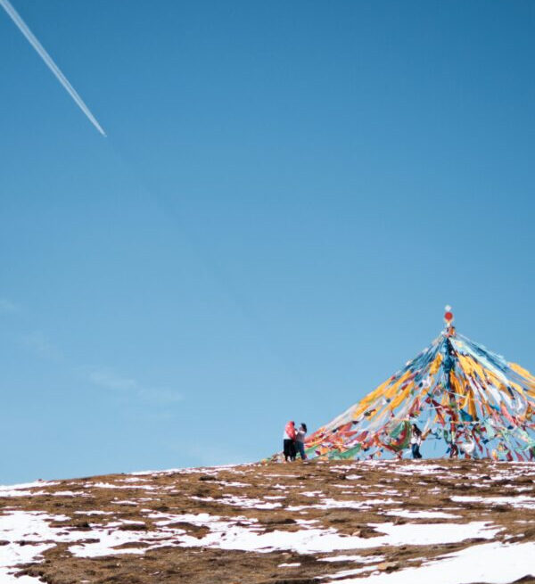 Two people stand near a colorful prayer flag pyramid on a snow-dusted hill against a clear blue sky.