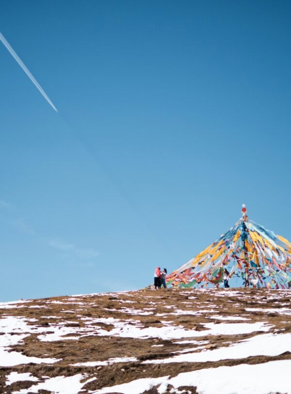 Dongda Mountain Pass,Tibet,Chinatour Two people stand near a colorful prayer flag pyramid on a snow-dusted hill against a clear blue sky.