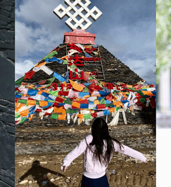 Girl in a white jacket and navy skirt stands with arms outstretched before a multi-colored prayer-flag covered stupa under a blue sky.