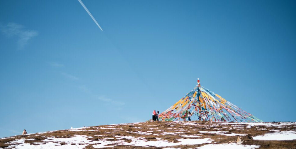 Blue sky over a snow-dusted hillside with a colorful conical prayer-flag structure and a few people nearby.
