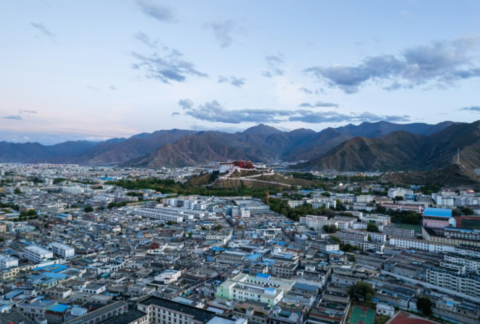 Tibet,Chinatour Aerial view of a densely built city with a hilltop fortress or structure and distant mountains under a clear blue sky.