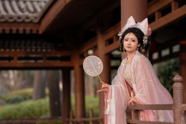 Hanfu,China Culture,Chinatour Young woman in pink traditional Hanfu with ornate headpiece, standing on a wooden temple veranda holding a decorative round fan.