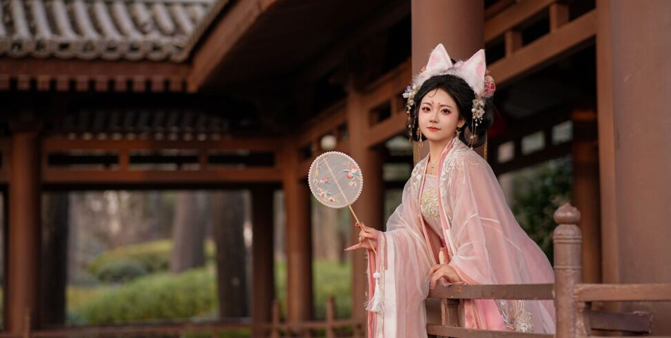 Young woman in pink traditional Hanfu with ornate headpiece, standing on a wooden temple veranda holding a decorative round fan.