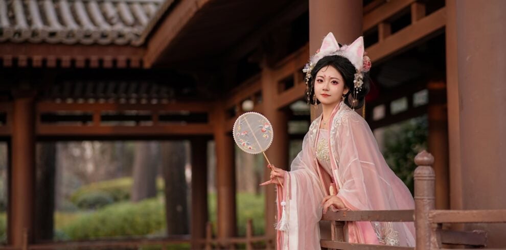 Hanfu,China Culture,Chinatour Young woman in pink traditional Hanfu with ornate headpiece, standing on a wooden temple veranda holding a decorative round fan.