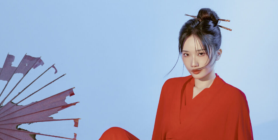Portrait of a young woman in a red outfit against a light blue background, with a pink fan visible on the left side.