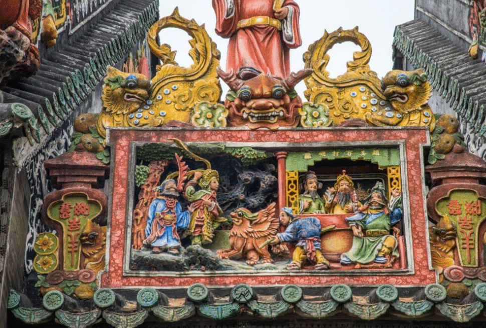 Colorful carved temple panel with figures around a central dragon and mythic creatures, ornate green tiled roof edges of a traditional roof.