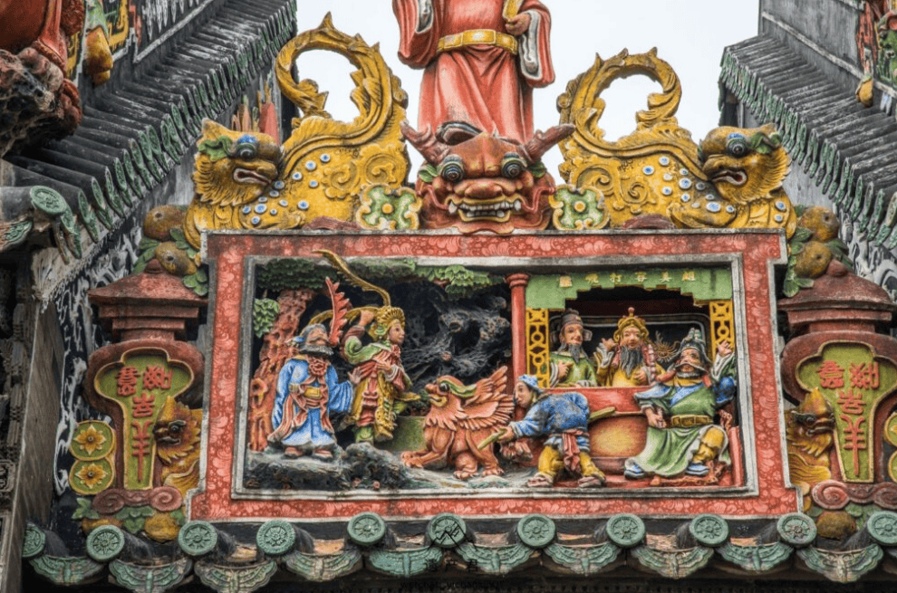 Colorful carved temple panel with figures around a central dragon and mythic creatures, ornate green tiled roof edges of a traditional roof.