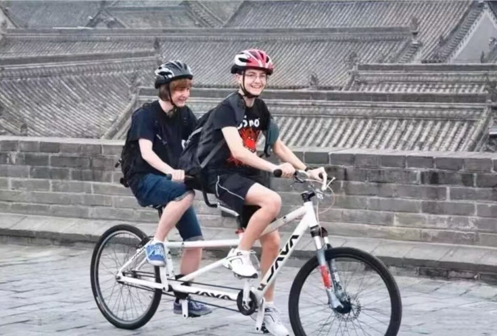 Xi'an Ancient City Wall: Cycling + Hanfu Photoshoots,Chinatour Two people ride a white tandem bicycle wearing helmets along a stone-paved street with a stone wall in the background (front rider smiling).