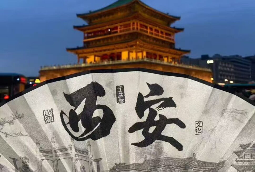 Xi'an Muslim Quarter Food,Chinatour Night scene with a lit multi-tier Chinese pagoda behind a large decorative fan featuring bold calligraphy and ink drawings in the foreground.