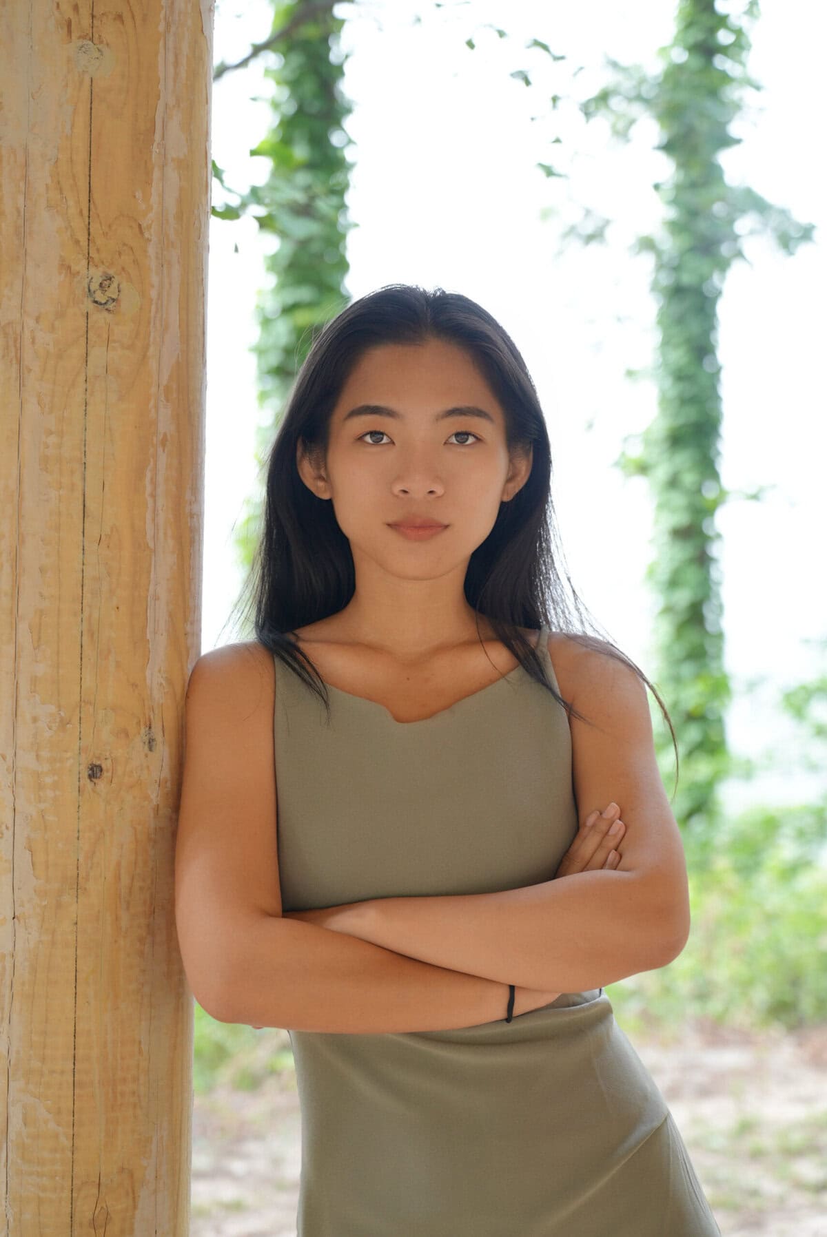 Lesley Chen, China Explorer Tour Team, chinatravel, Young woman stands outdoors, arms crossed, leaning against a wooden post with greenery in the background.