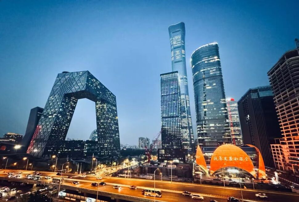 Night cityscape of a modern business district with a diamond-patterned arch-shaped building and glowing orange pavilion by a busy highway at dusk/lighted skyscrapers nearby