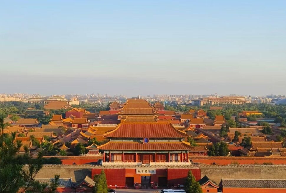 Panoramic view of the Forbidden City in Beijing, with red walls, golden-tiled roofs, and a distant city skyline on the horizon.