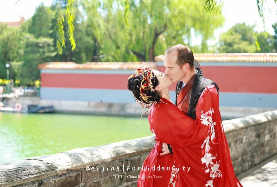Memorable Couple Hanfu Photoshoot,Chinatour Couple in red traditional Chinese outfits kissing by a stone railing along a green waterway, with park in the background.