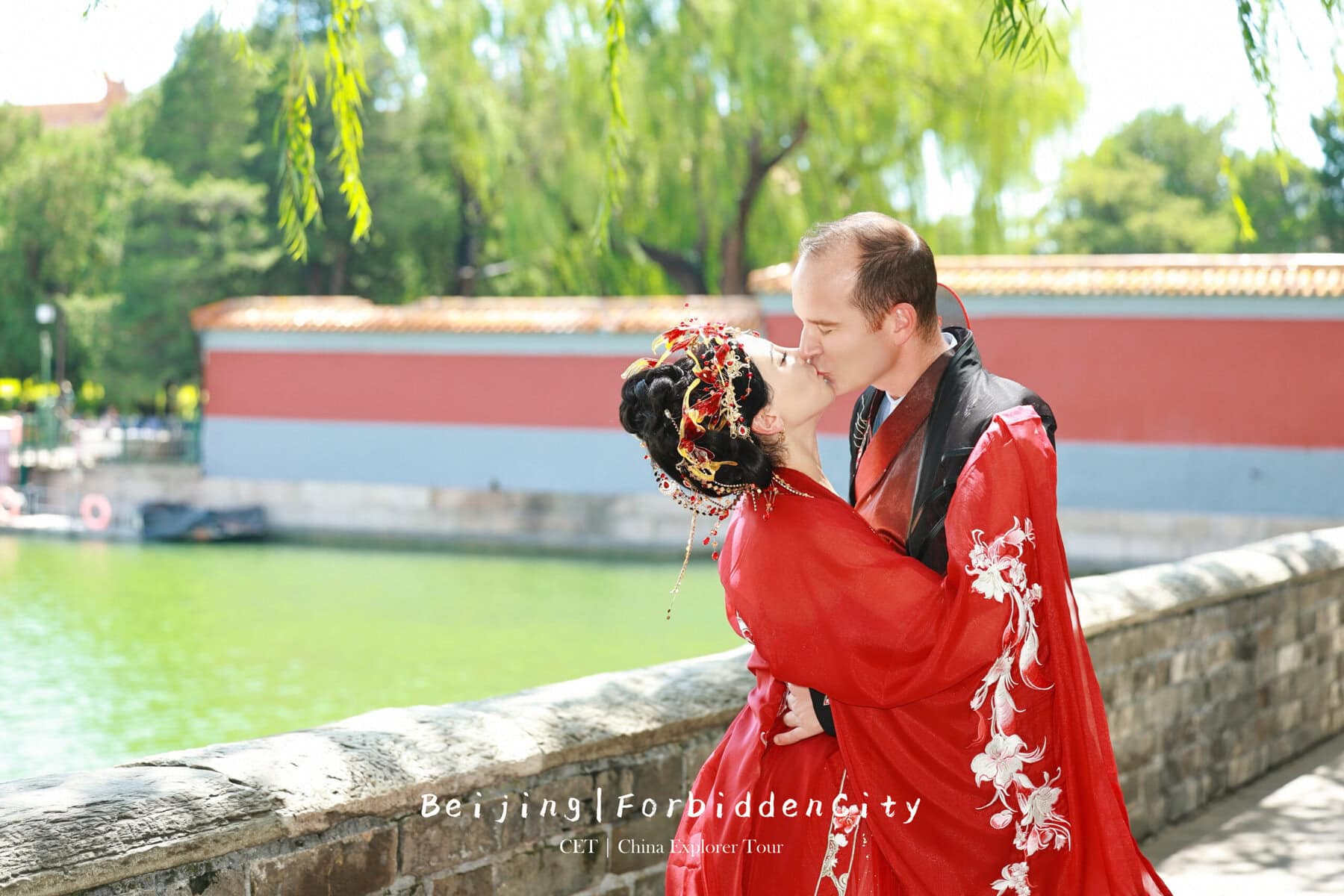 Memorable Couple Hanfu Photoshoot,Chinatour Couple in red traditional Chinese outfits kissing by a stone railing along a green waterway, with park in the background.