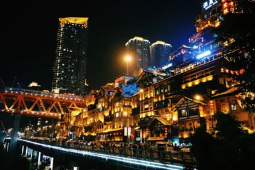 Night view of a brightly lit riverside street with traditional-style wooden buildings, a glowing bridge, and tall buildings in the background reflected on the river at night.