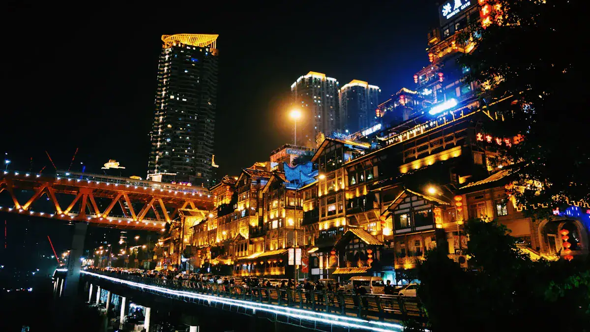 Night view of a brightly lit riverside street with traditional-style wooden buildings, a glowing bridge, and tall buildings in the background reflected on the river at night.