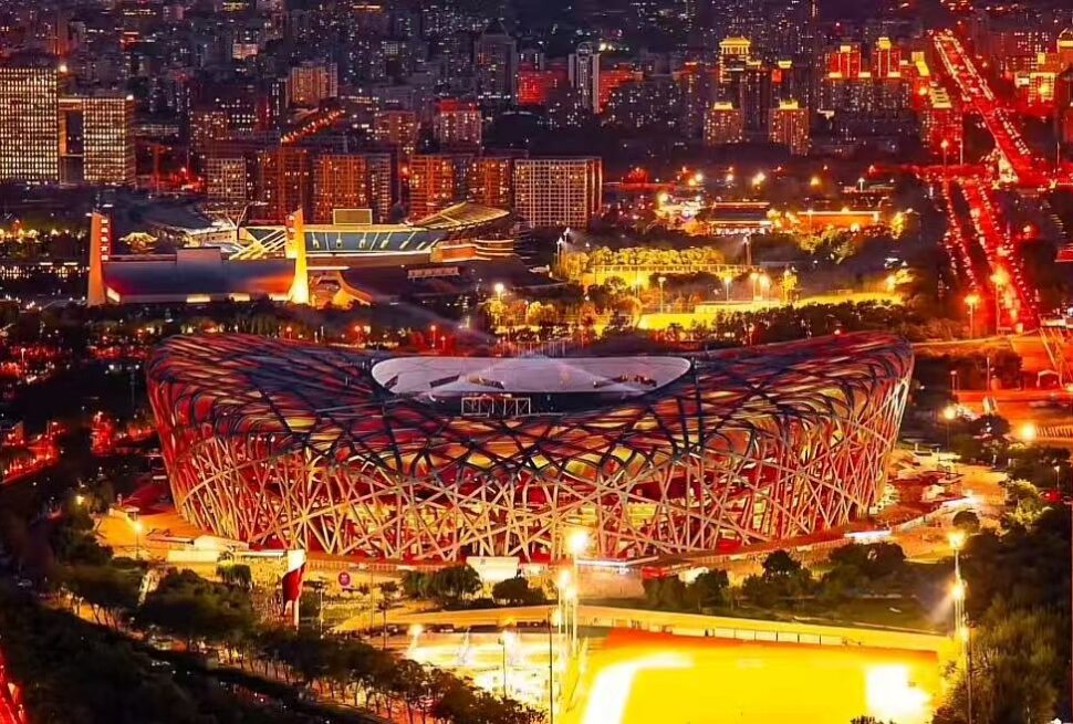 Night aerial view of the Beijing National Stadium (Bird's Nest) illuminated in red and orange, with city lights in the background.