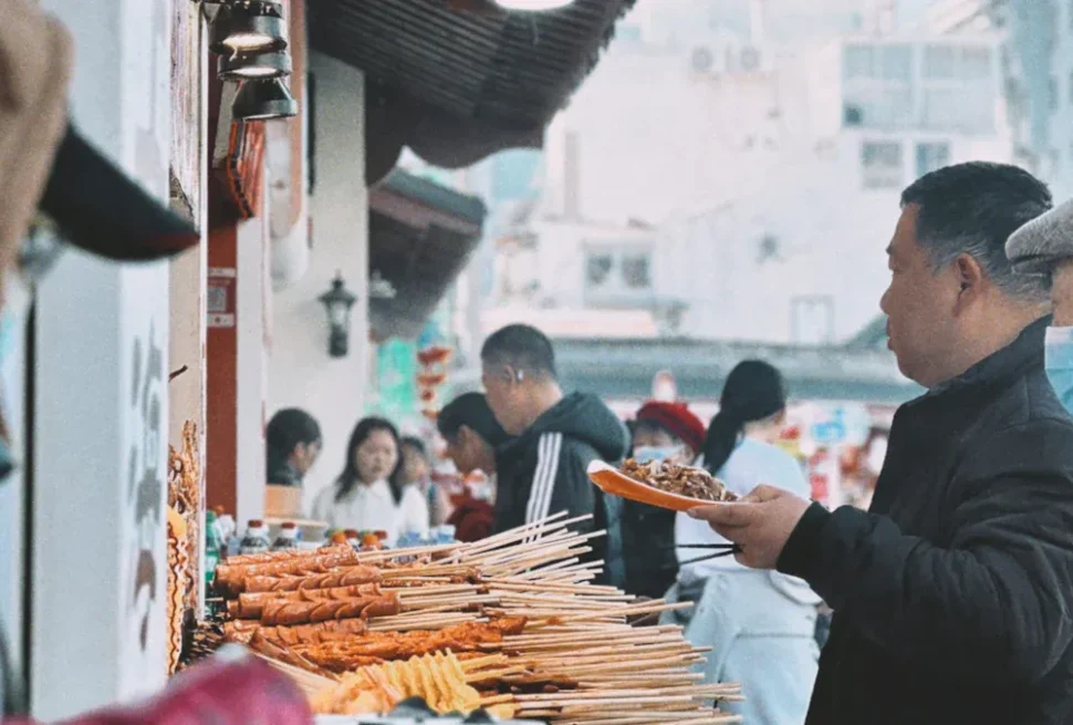 People at a street food stall with skewered kebabs and noodles on display, someone serving meat on a plate while others shop nearby.