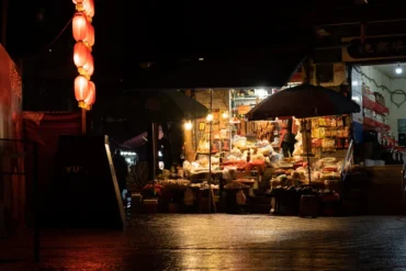 Night market stall at night under red lanterns, with goods displayed and shoppers nearby under umbrellas.