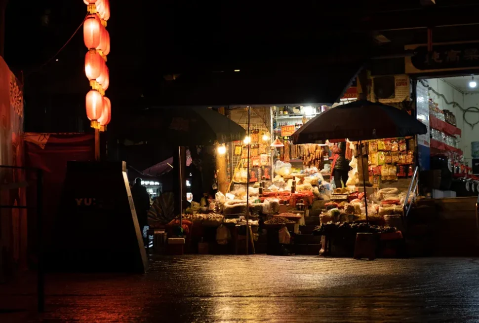 Night market stall at night under red lanterns, with goods displayed and shoppers nearby under umbrellas.