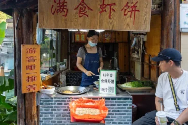 Woman wearing a face mask and blue apron cooks shrimp pancakes at an open-air street food stall; a customer sits nearby with a drink.