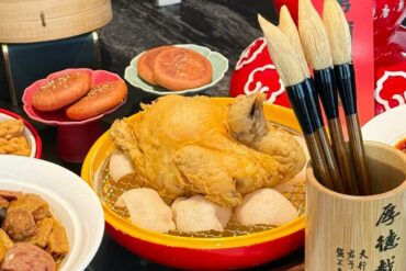 Festive Chinese banquet with a fried whole chicken in a yellow dish, buns, and a wooden brush holder with calligraphy on the right.