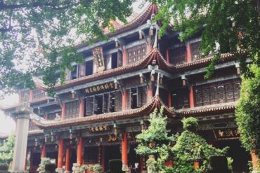 Multi-story Chinese temple with curved tiled roofs, red columns, gold plaques, and surrounding greenery.