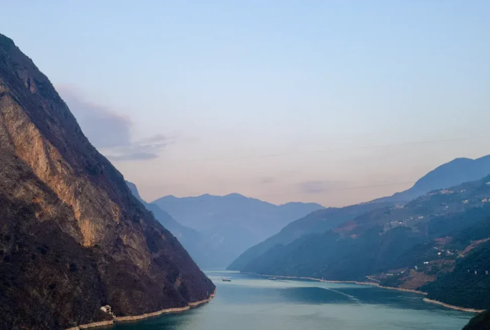 Wide river winds between steep, shadowed mountains at dusk with a pale blue sky above