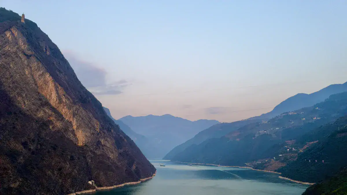 Wide river winds between steep, shadowed mountains at dusk with a pale blue sky above