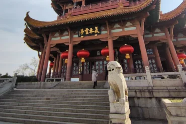 Chinese temple entrance with red lanterns, red pillars, and curved ornate roofs; a stone lion guards the stairs in front of the building.