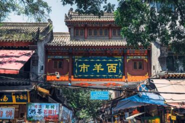 Decorative Chinese gateway over a bustling market street with shops and stalls beneath a blue sign panel and ornate roof.