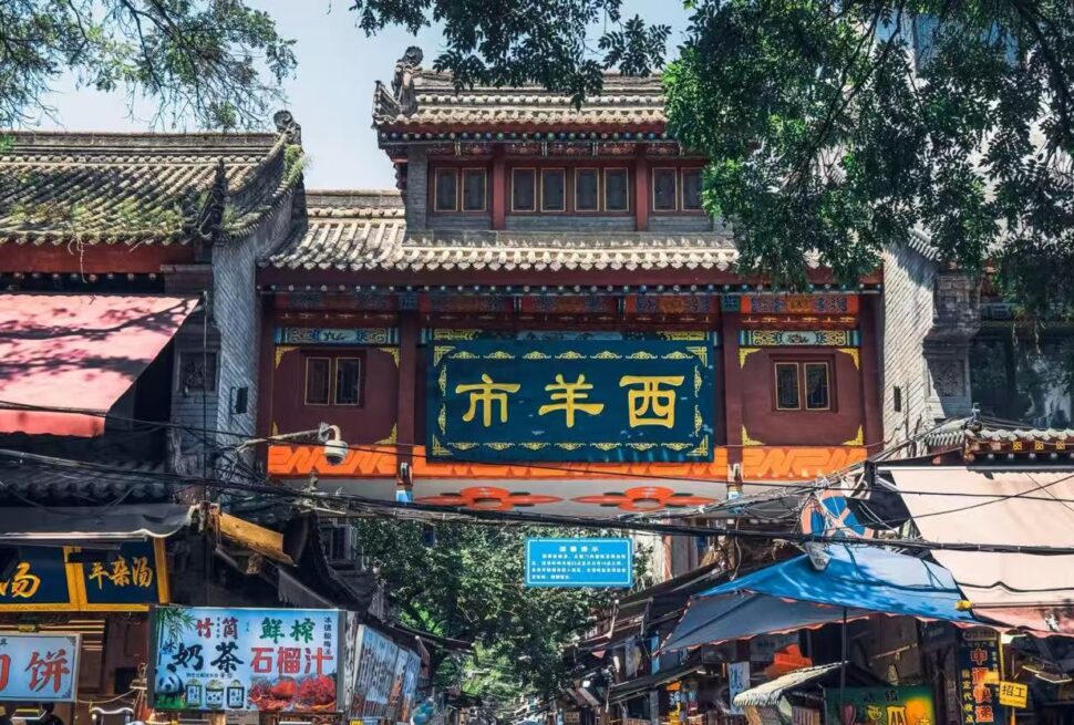 Xi'an Muslim Quarter,Chinatour Decorative Chinese gateway over a bustling market street with shops and stalls beneath a blue sign panel and ornate roof.